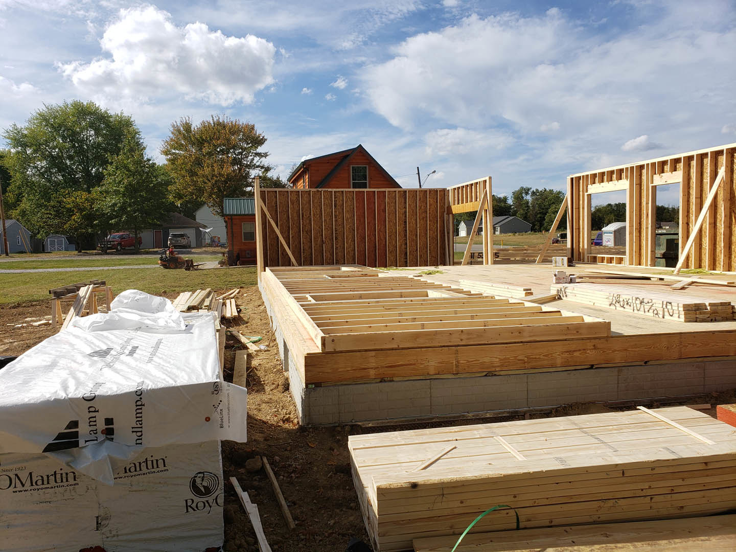 Wood framing and beams on a partially constructed house, stacks of lumber and fencing in foreground, mature trees and blue sky with scattered clouds in background