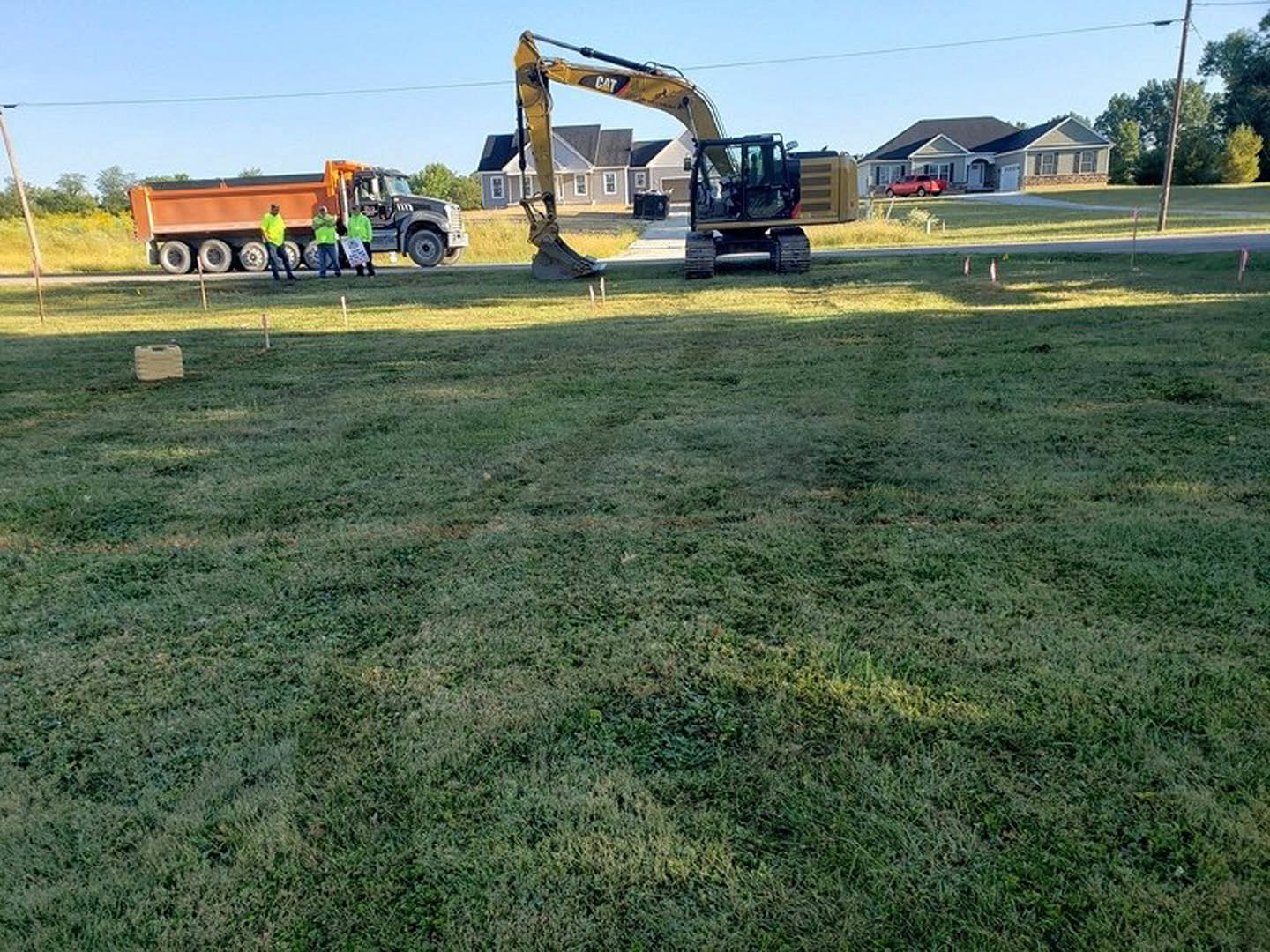 Construction site with grassy field, yellow-shirted workers, tractor and truck parked near dirt lot, trees and open sky in background