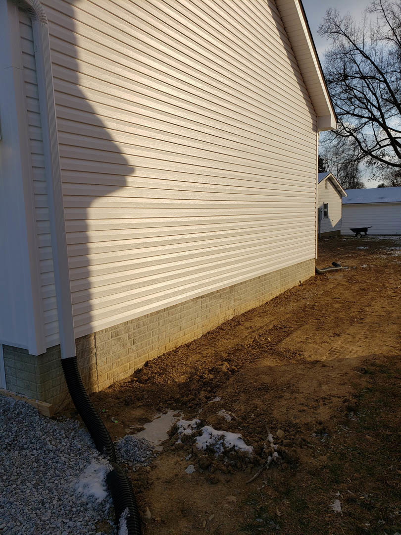 White siding exterior with black downspout, wheelbarrow on dirt patch, tree beside house, visible roof and windows