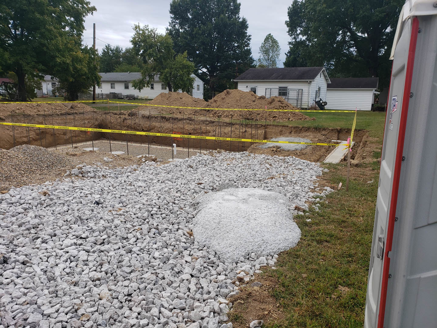Gravel-covered construction site with a pile of white rocks, dirt mound beside a white house, yellow caution tape, and portable toilet in the foreground