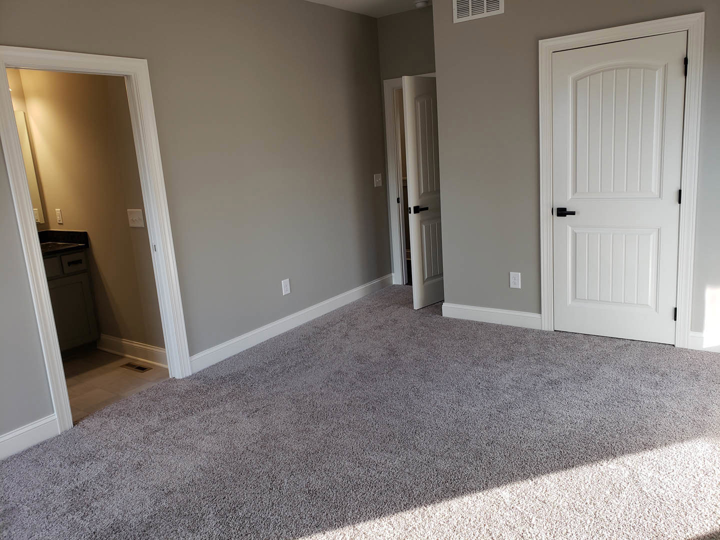 Carpeted room with white paneled doors, black handles, white trim, open doorway leading to bathroom with sink and mirror, close-up of floor vent