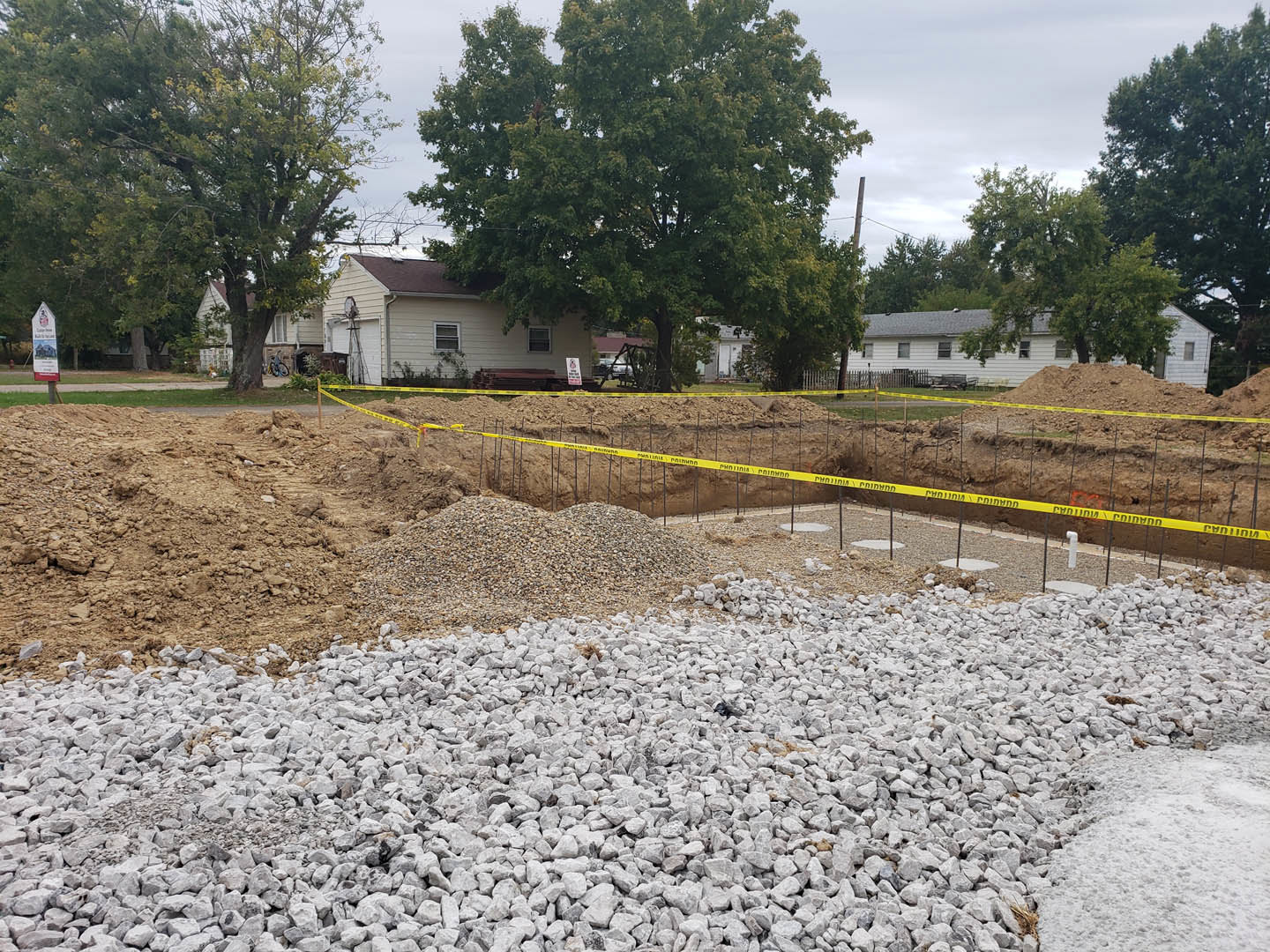Gravel and rocks piled beside a tree with leafy branches, yellow caution tape encircling a hole in the ground near a partially visible house exterior