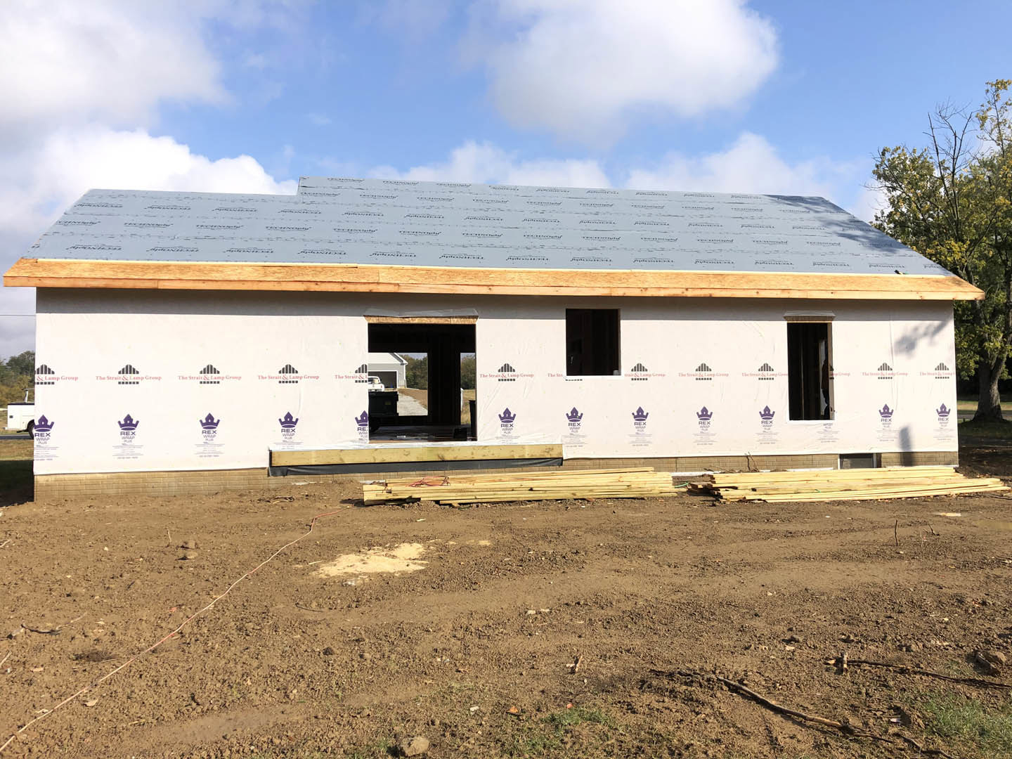 Partially built house with exposed wooden framing, shingled roof, dirt yard, scattered lumber, and a person standing in the doorway
