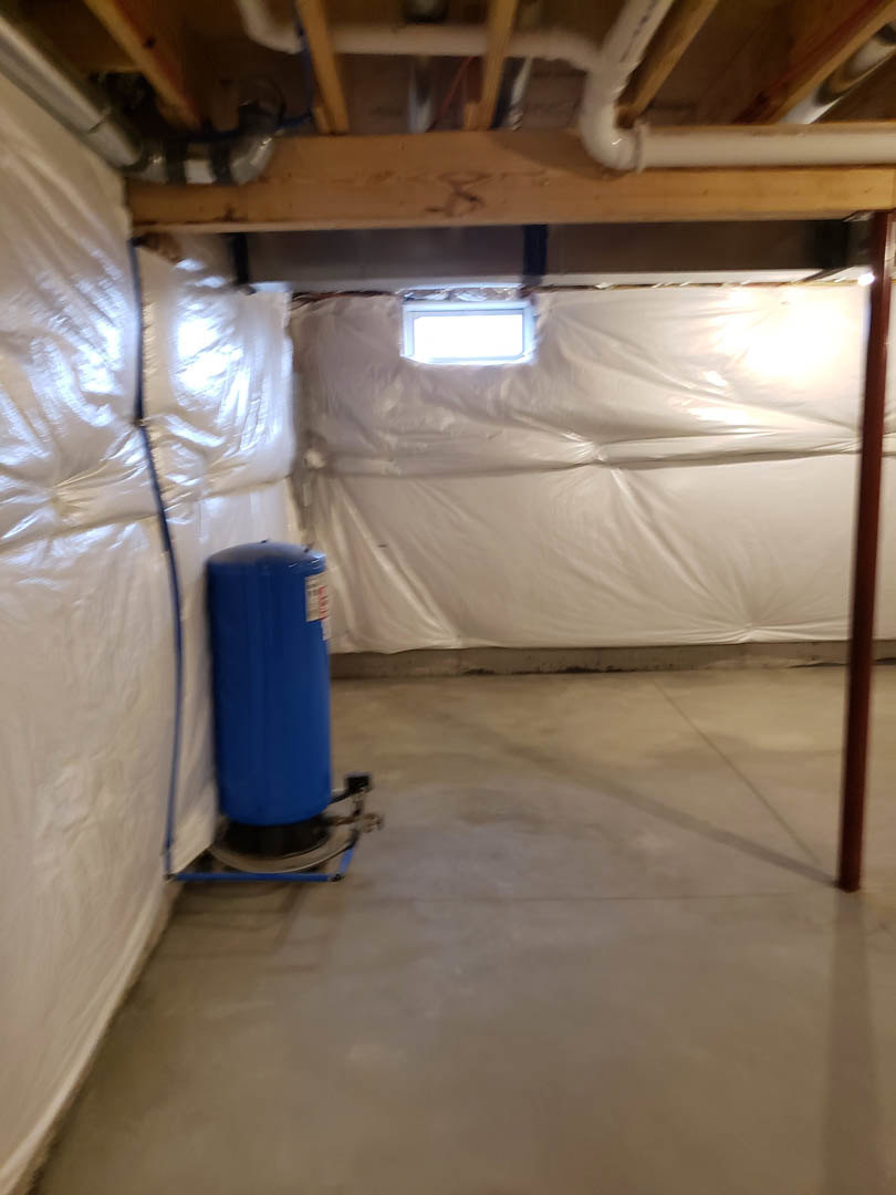 Blue water heater with white label standing on concrete basement floor, plastic sheet covering wall, exposed ceiling beams visible above.