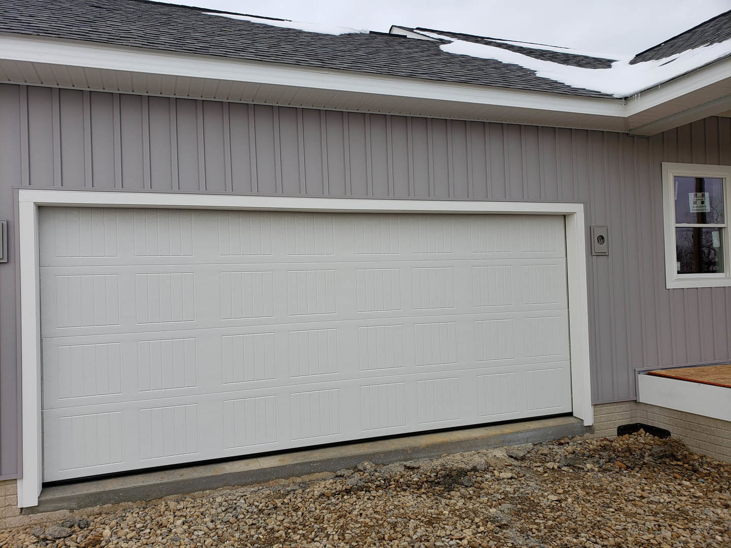 Gray paneled garage door beneath matching roof, stone pile along driveway, window with sign, light-colored siding exterior.