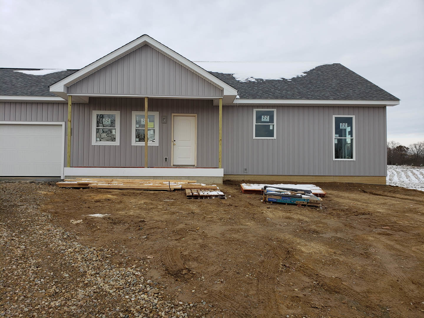 Light-colored siding home with white garage door, white entry door featuring gold handle, front window displaying a sign, and numerous wooden pallets stacked on dirt area in front.
