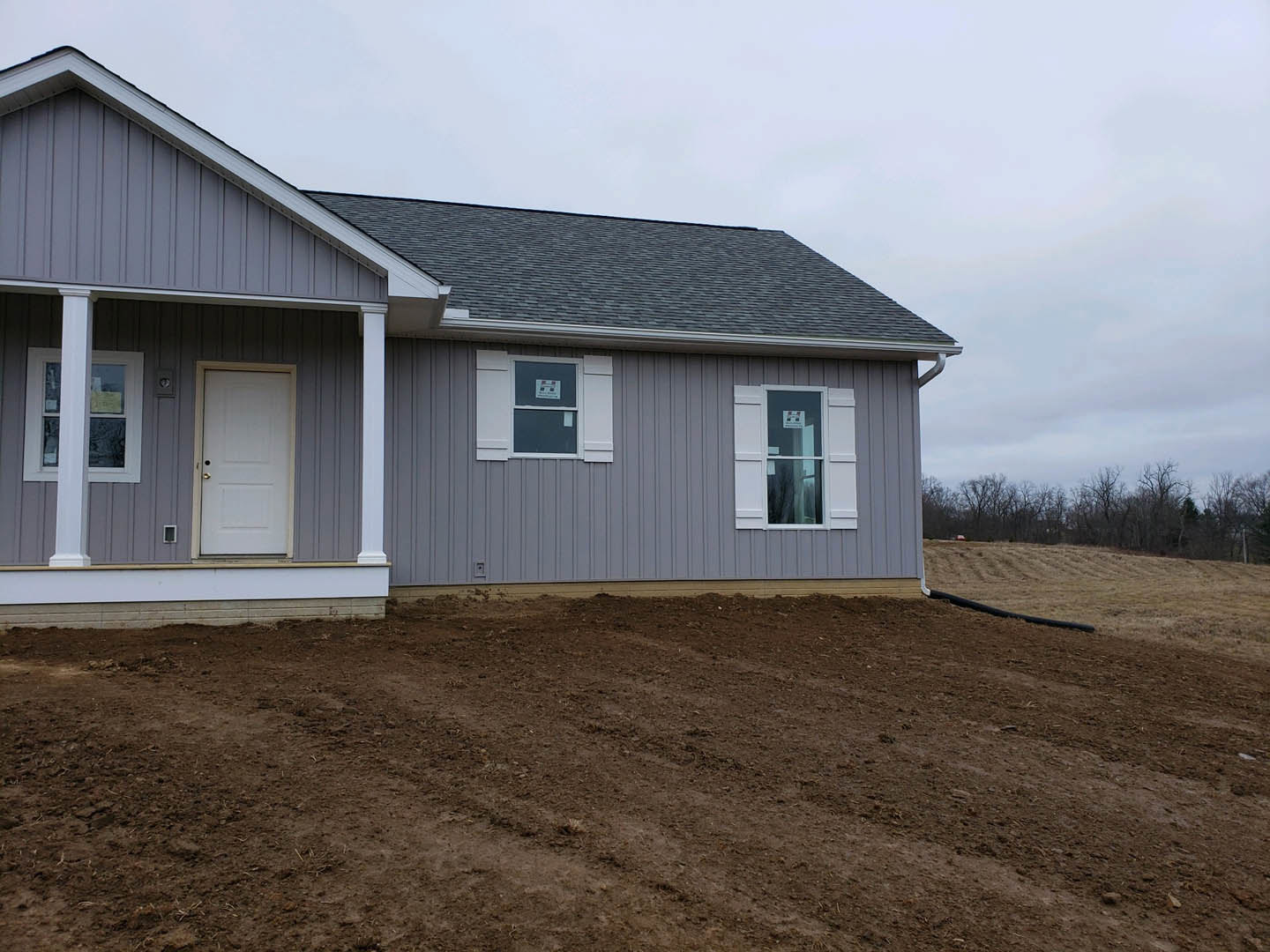 Single-story house with white siding, two white doors with gold handles, window displaying a sign, dirt field in foreground, white boundary wall, and group of trees in the