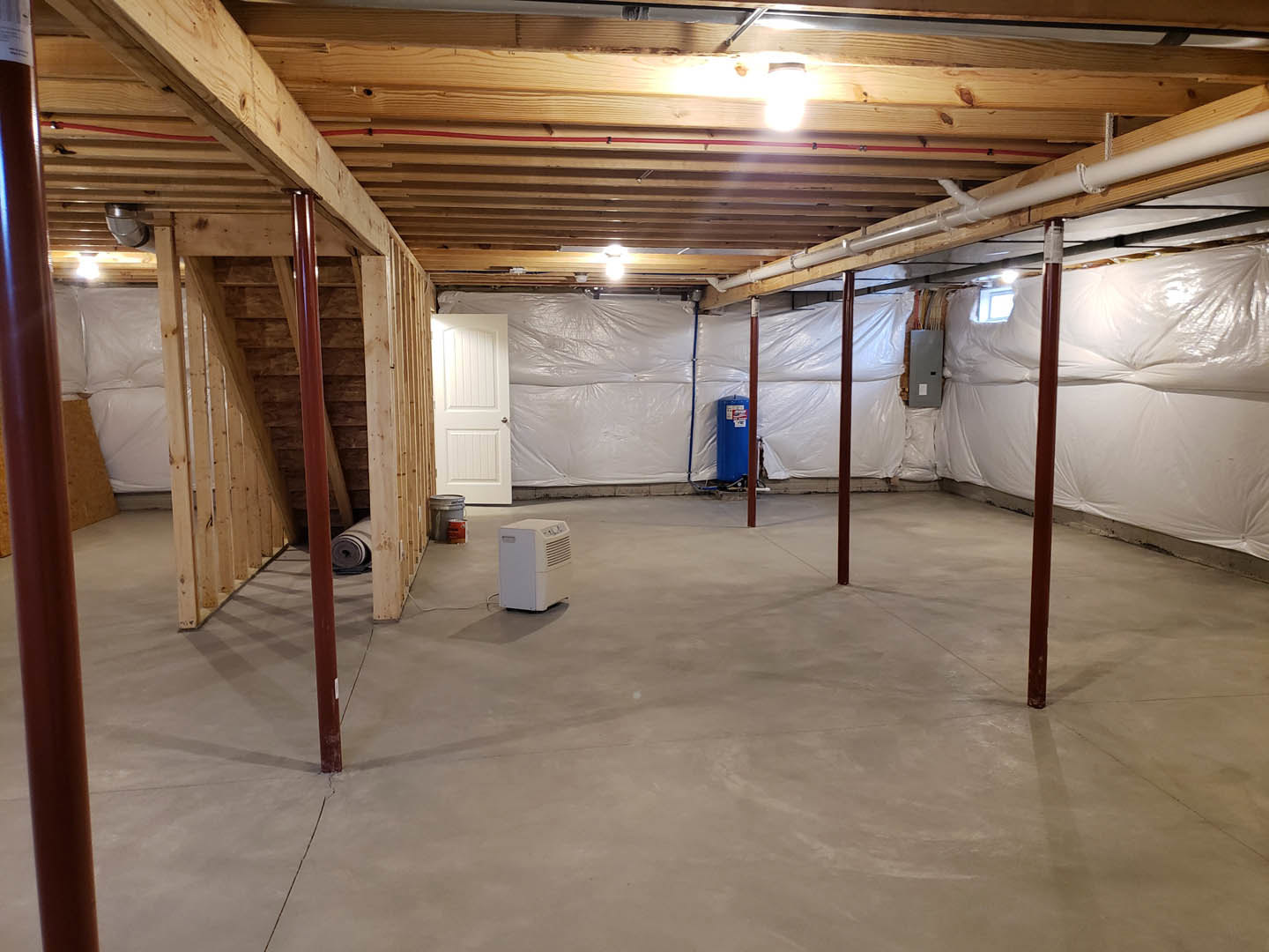 White-walled room with white ceiling, exposed concrete floor, white door with silver handle, white rectangular vent, roll of carpet on shelf, blue cylinder with red and white