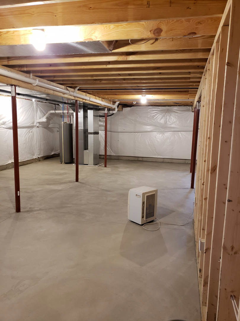 Unfinished basement with exposed ceiling beams, concrete floor, white rectangular HVAC vent box, and walls partially covered in white plastic insulation sheets