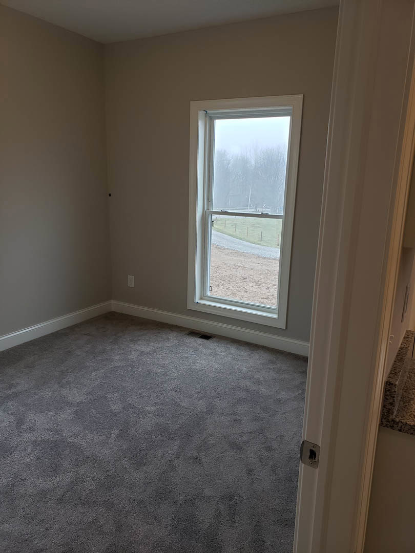 Carpeted room with white walls, large window overlooking road and trees, white door with glass panel, plaster ceiling