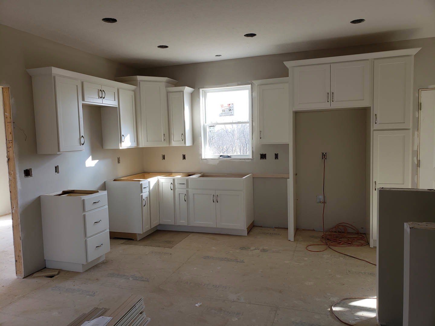 White kitchen cabinets, stainless refrigerator, window above sink, light countertops, wire on floor, missing drawer in lower cabinetry.