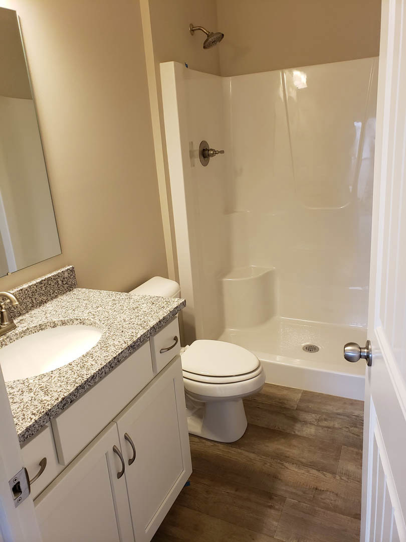 White porcelain sink and toilet on wood flooring, light-colored tile walls, chrome faucet, frameless mirror above countertop.