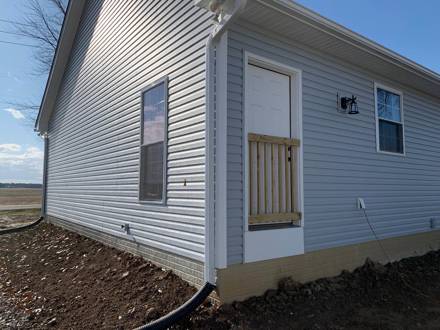 White front door with glass panel, black downspout, white siding, wooden railing, adjacent window, patch of bare dirt near entry.
