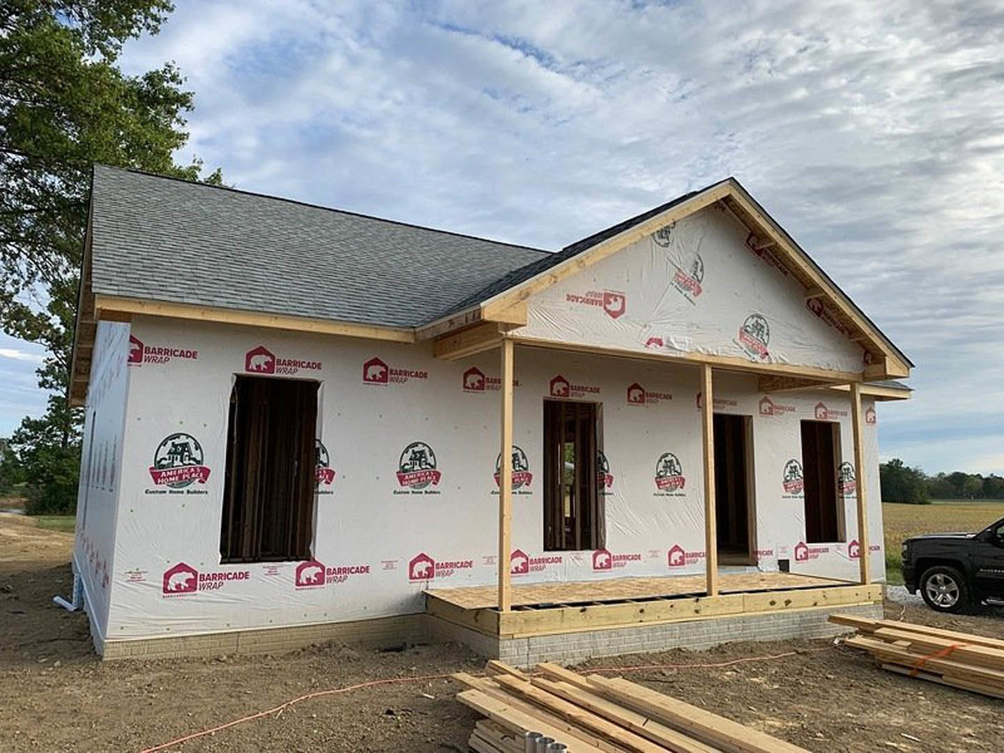 Wood-framed house under construction with covered porch, exposed beams, and construction materials scattered on dirt lot