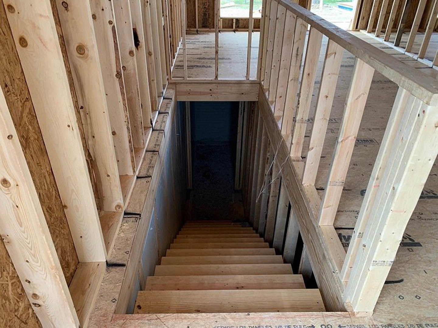 Wooden staircase with matching handrail and balusters leading down to a basement, surrounded by neutral walls and a closed door in a dimly lit interior space