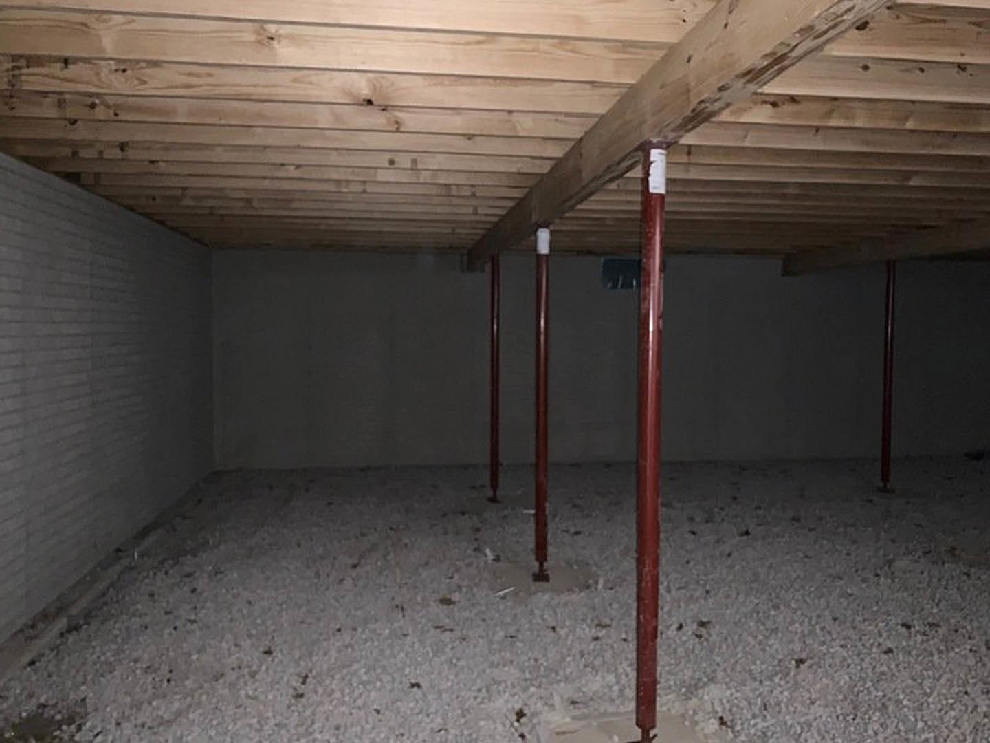 Basement with exposed wooden ceiling, red metal support poles, white gravel floor, and a window in the background.