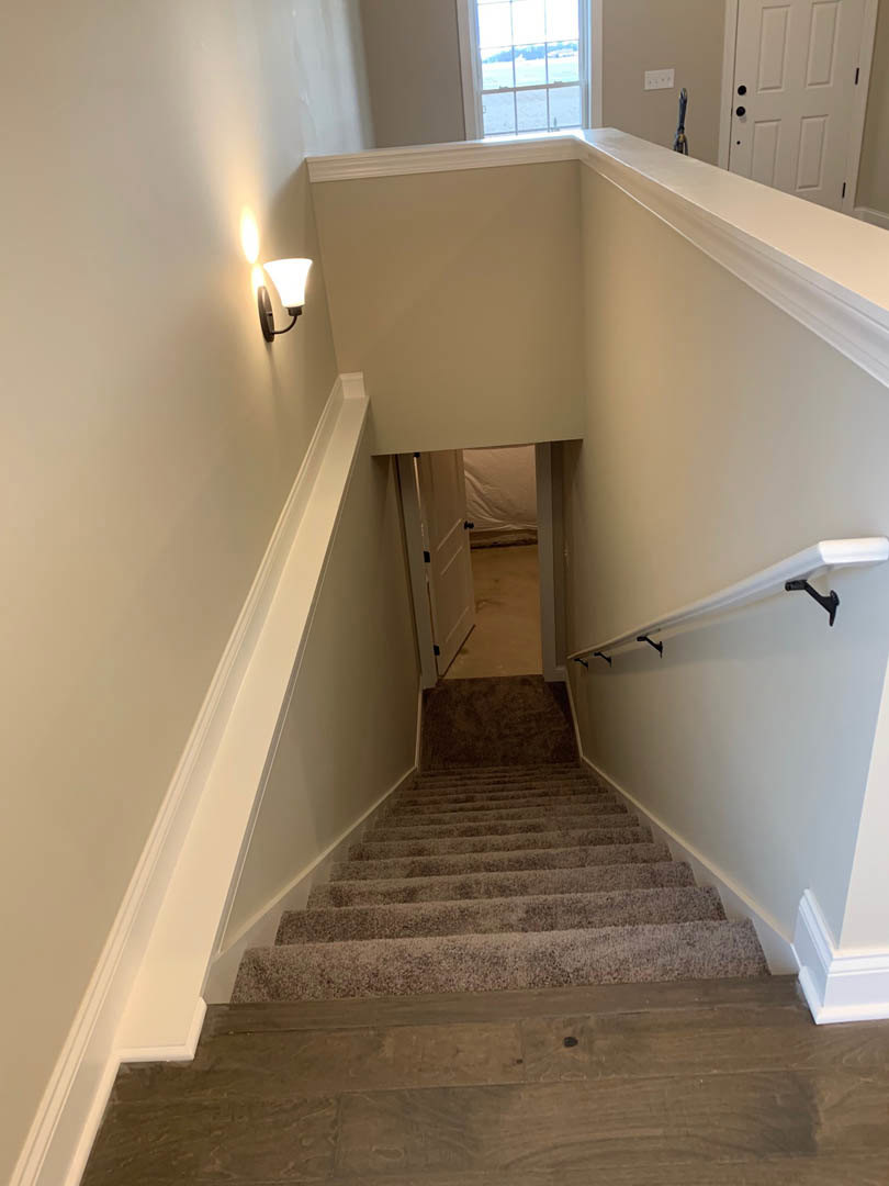 Wood staircase with plaster wall, warm ceiling light, and window overlooking grassy field; white curtain rod and lamp visible, hardwood flooring throughout.