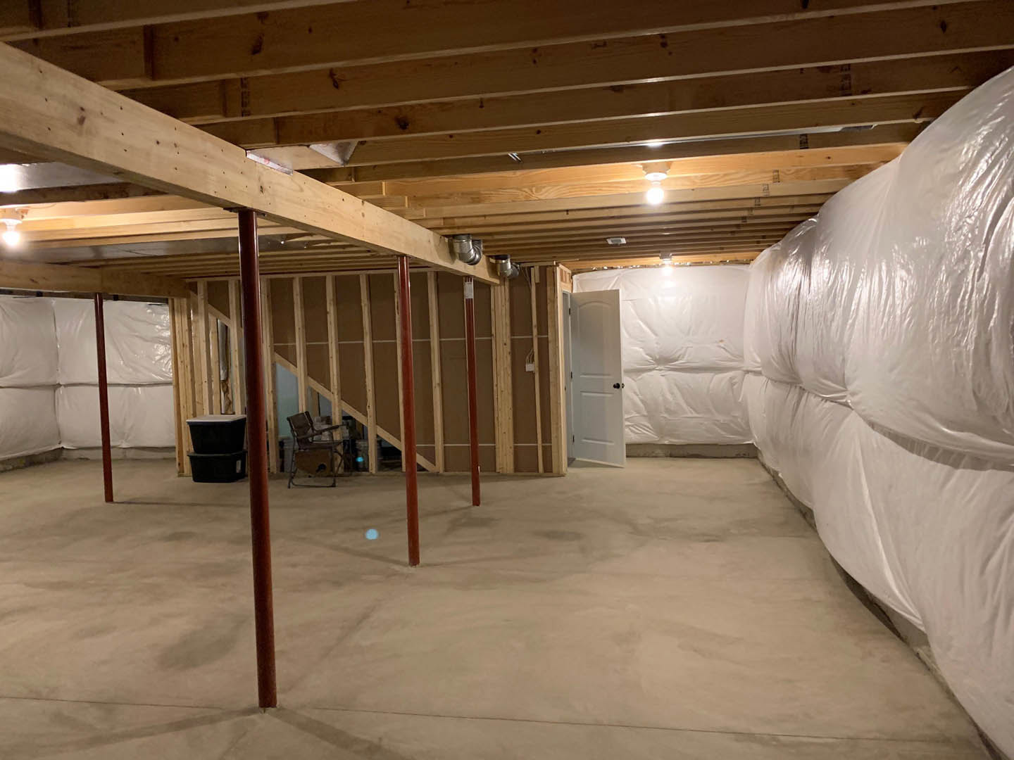 Basement with white plaster walls, white plastic sheet covering part of the space, white door with black knobs, stack of black plastic containers, chair placed on a table, white