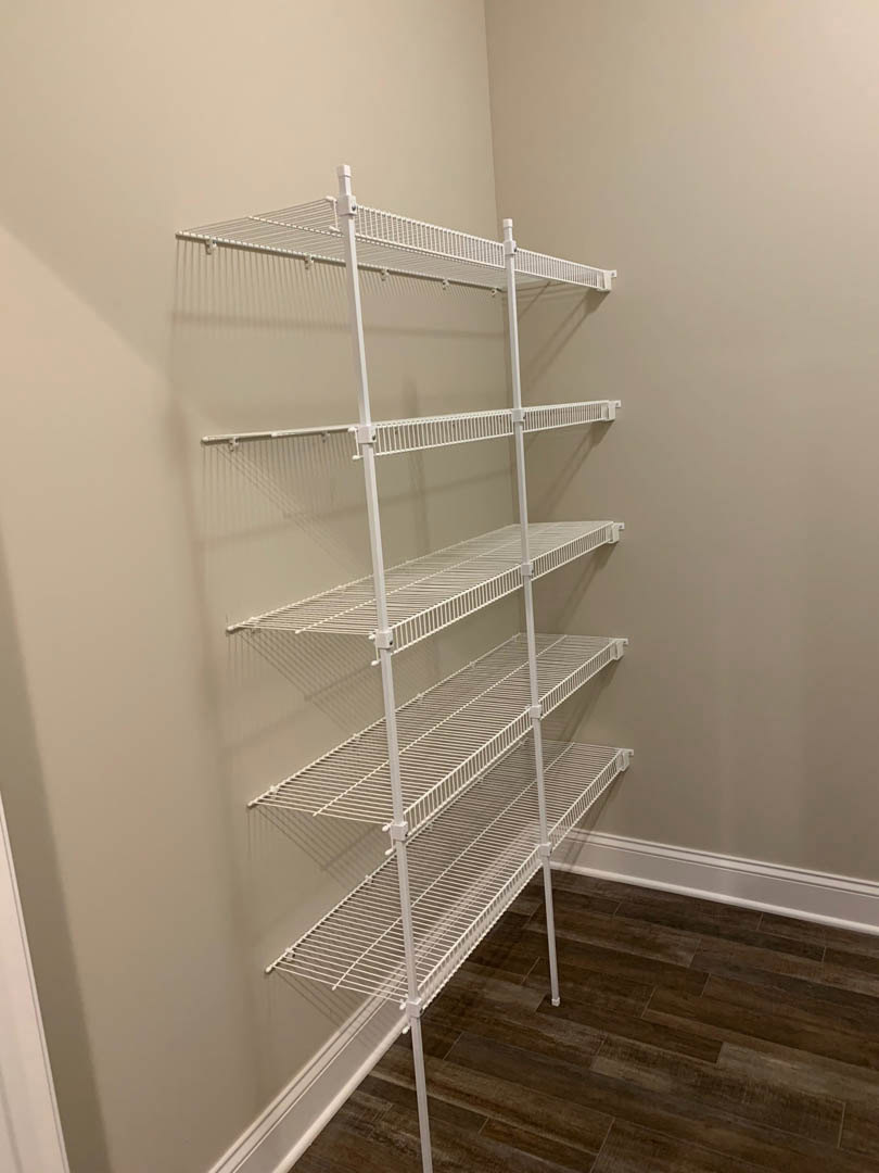 White wire shelving unit mounted on a white wall above wood flooring, featuring a metal rod and casting a subtle shadow.