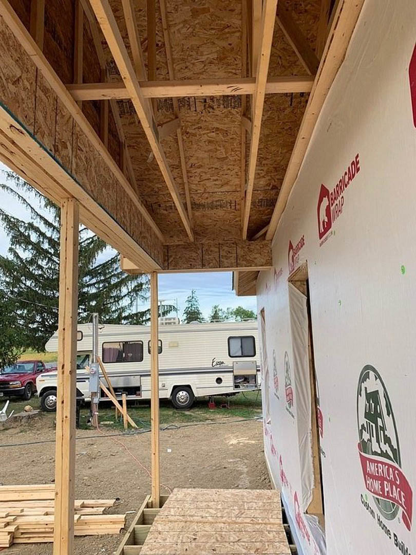 Partially built residential structure with exposed framing, white camper trailer parked nearby, mature tree in foreground, red truck and white car parked on dirt ground, scattered
