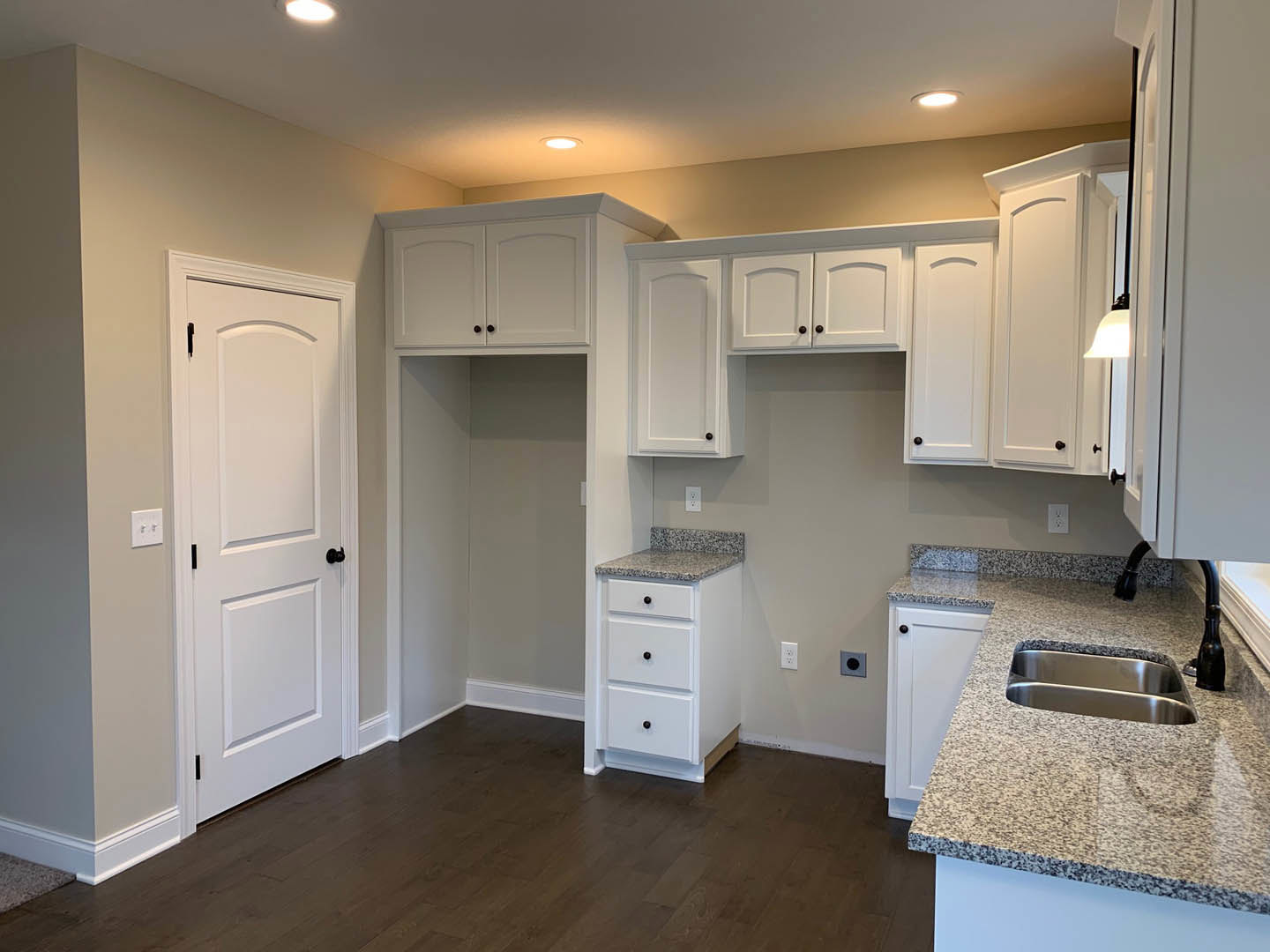 White kitchen cabinets with black knobs, granite countertops featuring two stainless steel sinks, white door with black knob, and light-colored flooring.