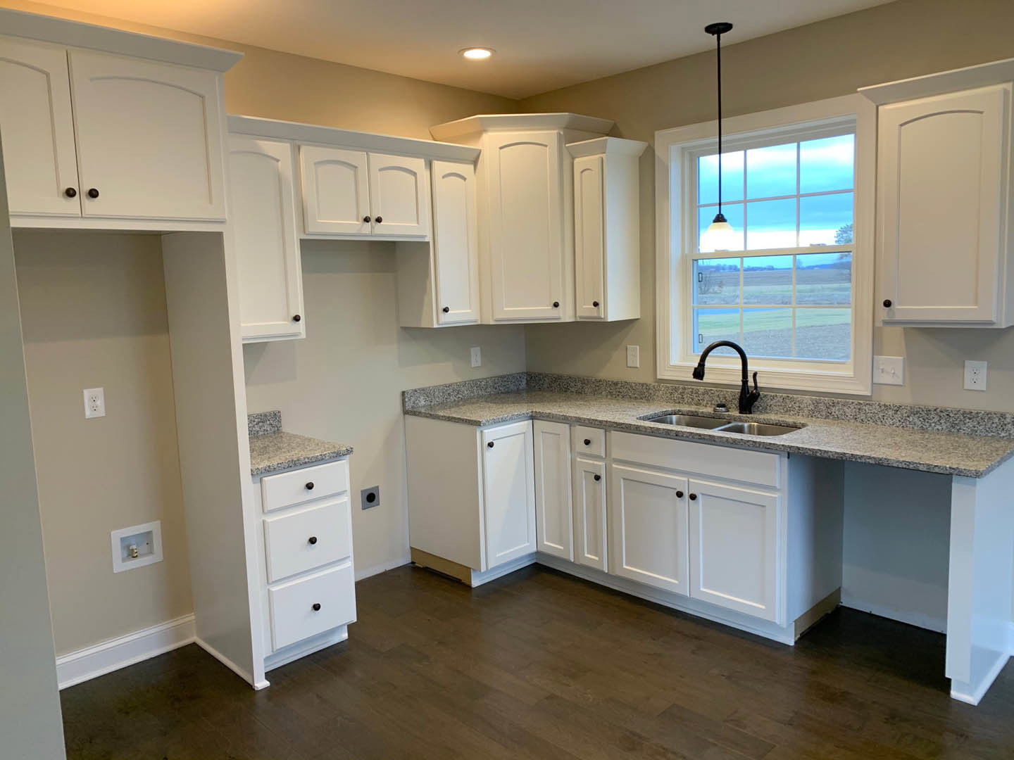 White kitchen cabinets and granite countertops, black faucet over stainless sink, window with ceiling light, white door with matching frame, dark wood flooring, white walls with