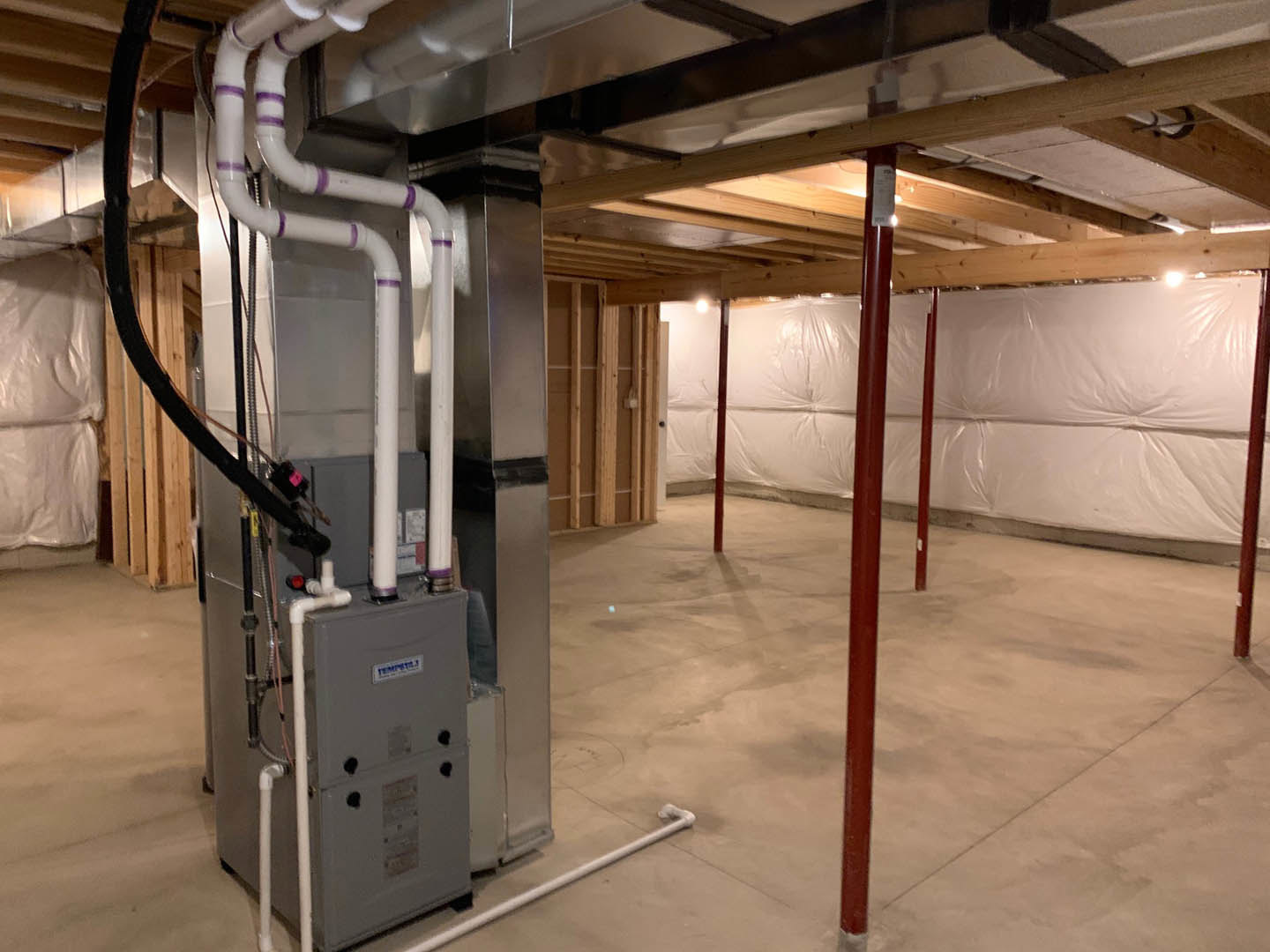 Exposed pipes and wires along ceiling and walls, white plastic sheeting on wall, grey electrical box, red metal poles, concrete floor