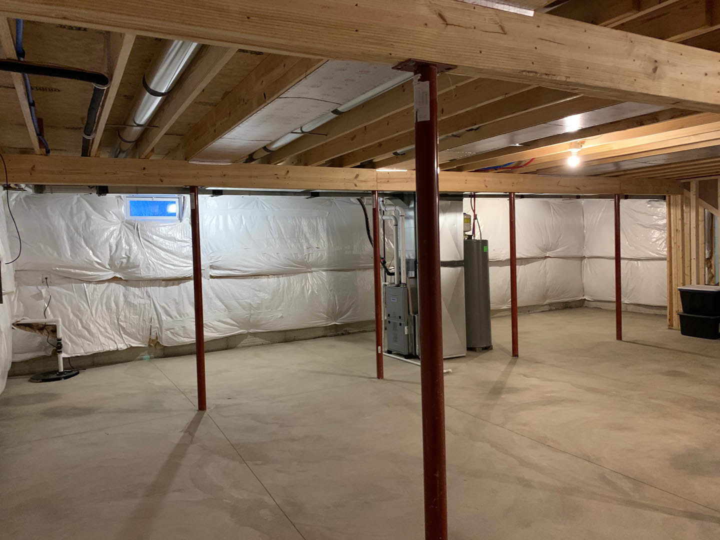 Basement with concrete floor, red support pole, white wall, exposed wooden beam with pipe, grey metal box with black holes, blue screen with light, and white sheet draped over