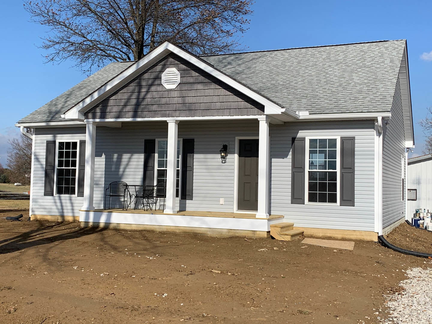 Gray-sided house with white-trimmed windows, covered porch, leafless tree, and bare dirt yard