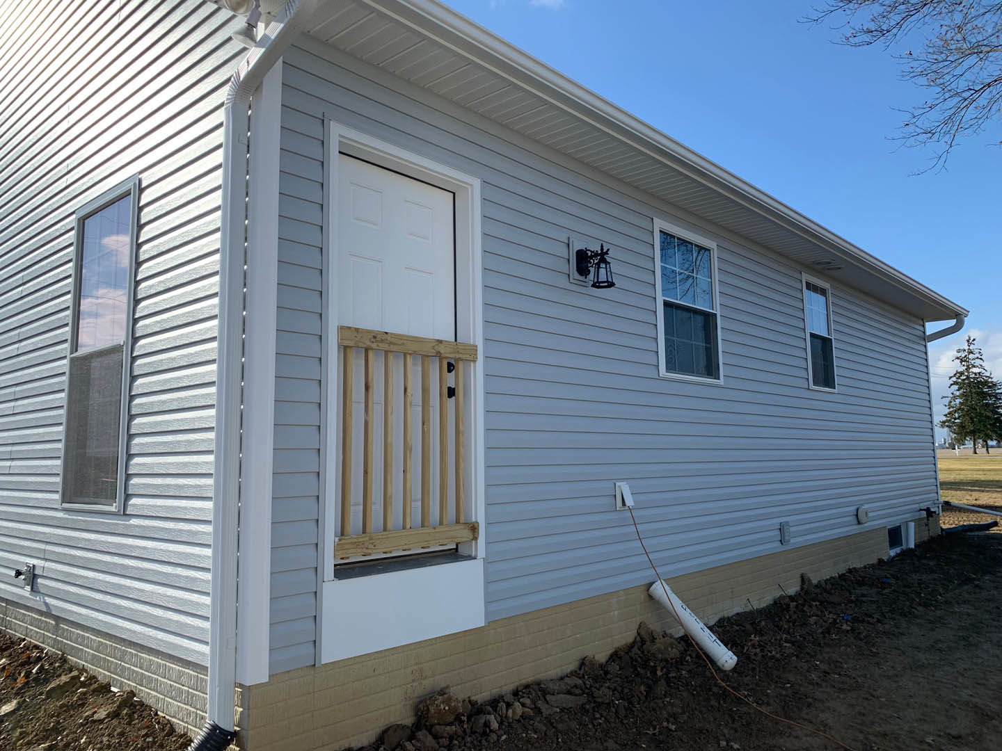 White siding exterior with wooden railing, single window reflecting tree branches, entry door, and exposed pipe; mature tree nearby.