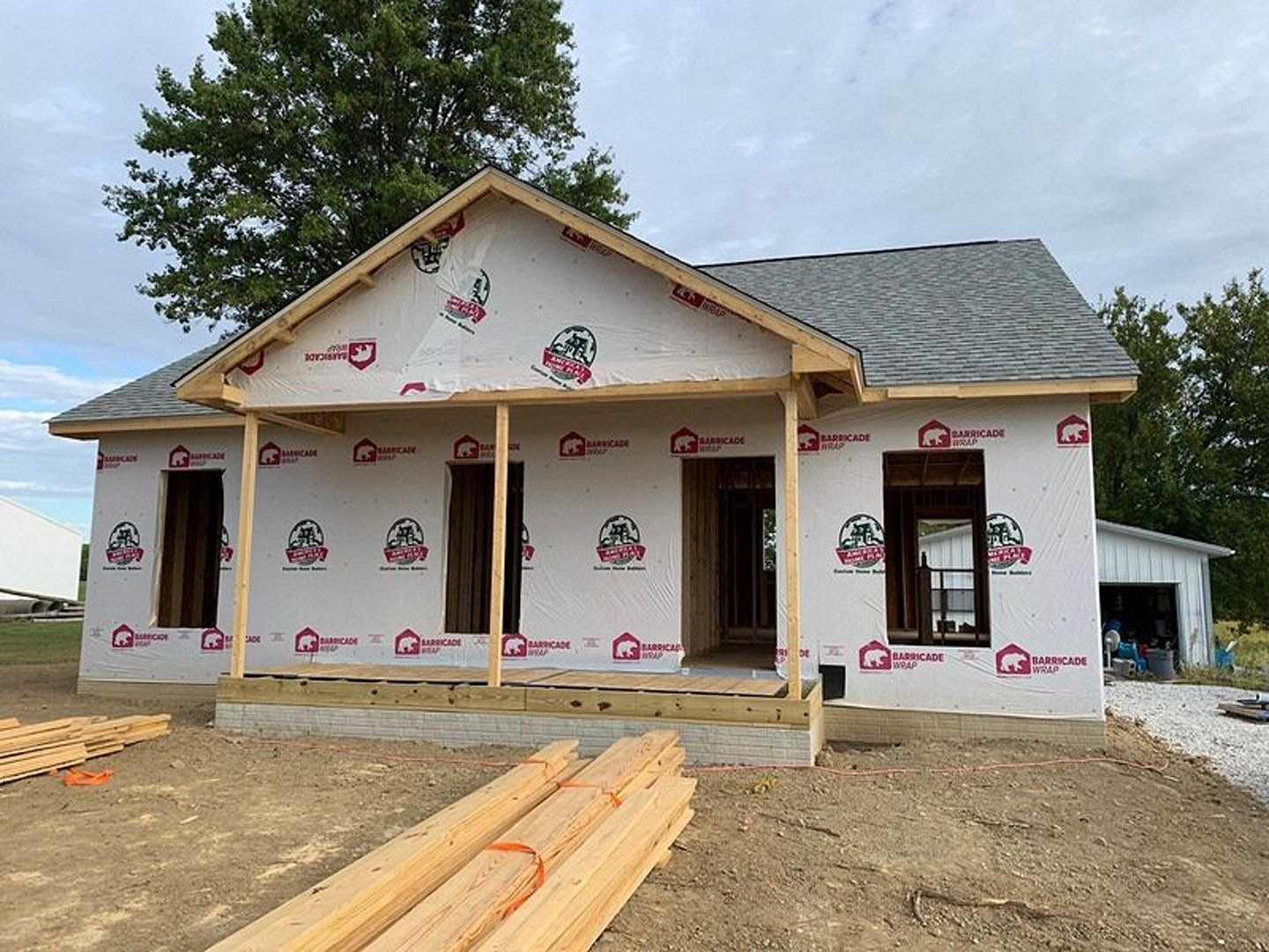 Framed house under construction with exposed wood beams, white sheet draped across front, pile of lumber on ground, unfinished doorway, roofline partially visible with tree and