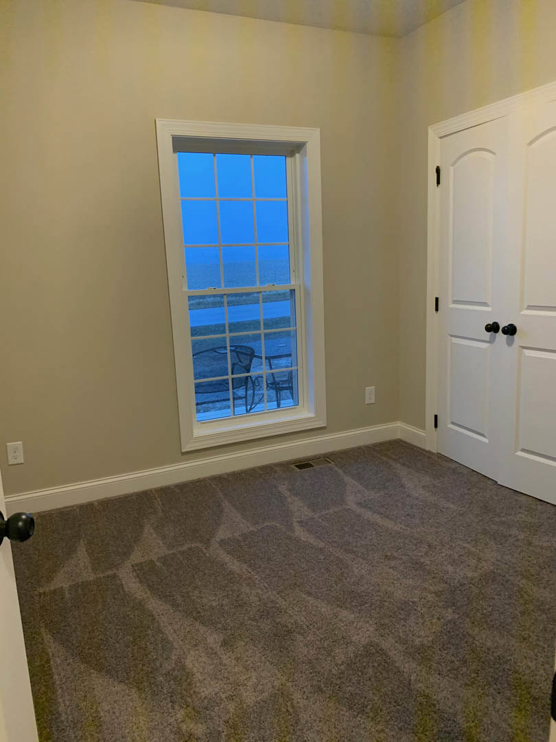 Carpeted bedroom with white walls, large window overlooking ocean, white door with black hardware, floor vent, and window featuring a small drawing.