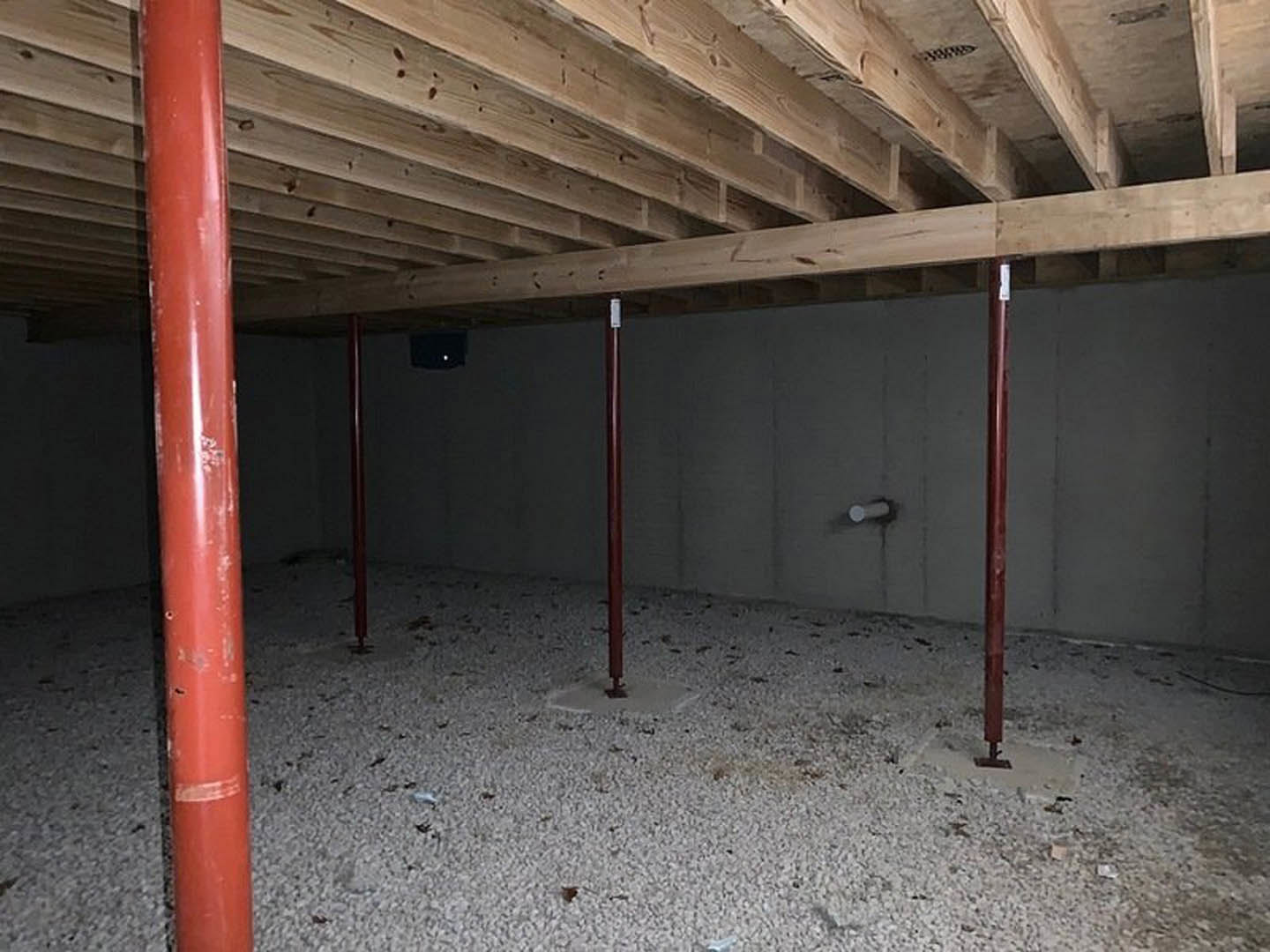 Exposed wooden ceiling beams above unfinished basement floor, red metal support pole near insulated walls, single overhead light illuminating space