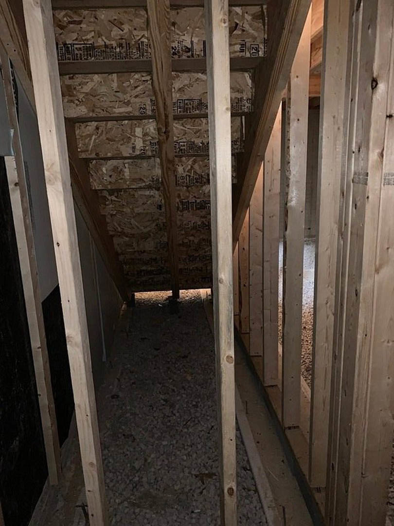 Exposed wooden beams and framing in an unfinished room, gravel scattered near the doorway, raw lumber and planks visible along walls.
