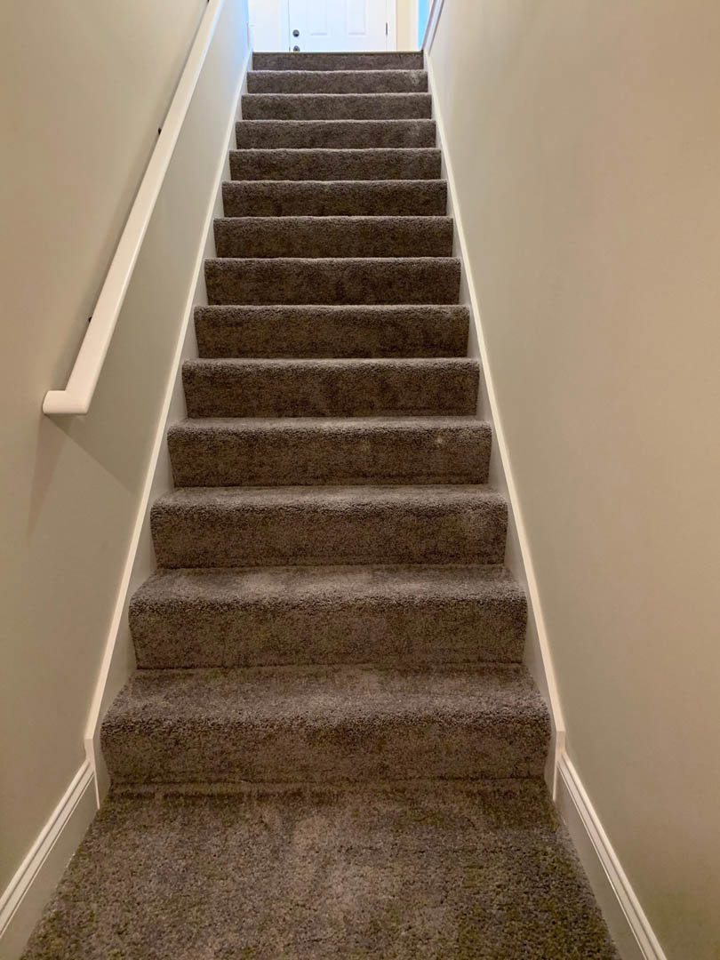 Carpeted staircase with wooden handrail, white walls, and a closed white door at the top