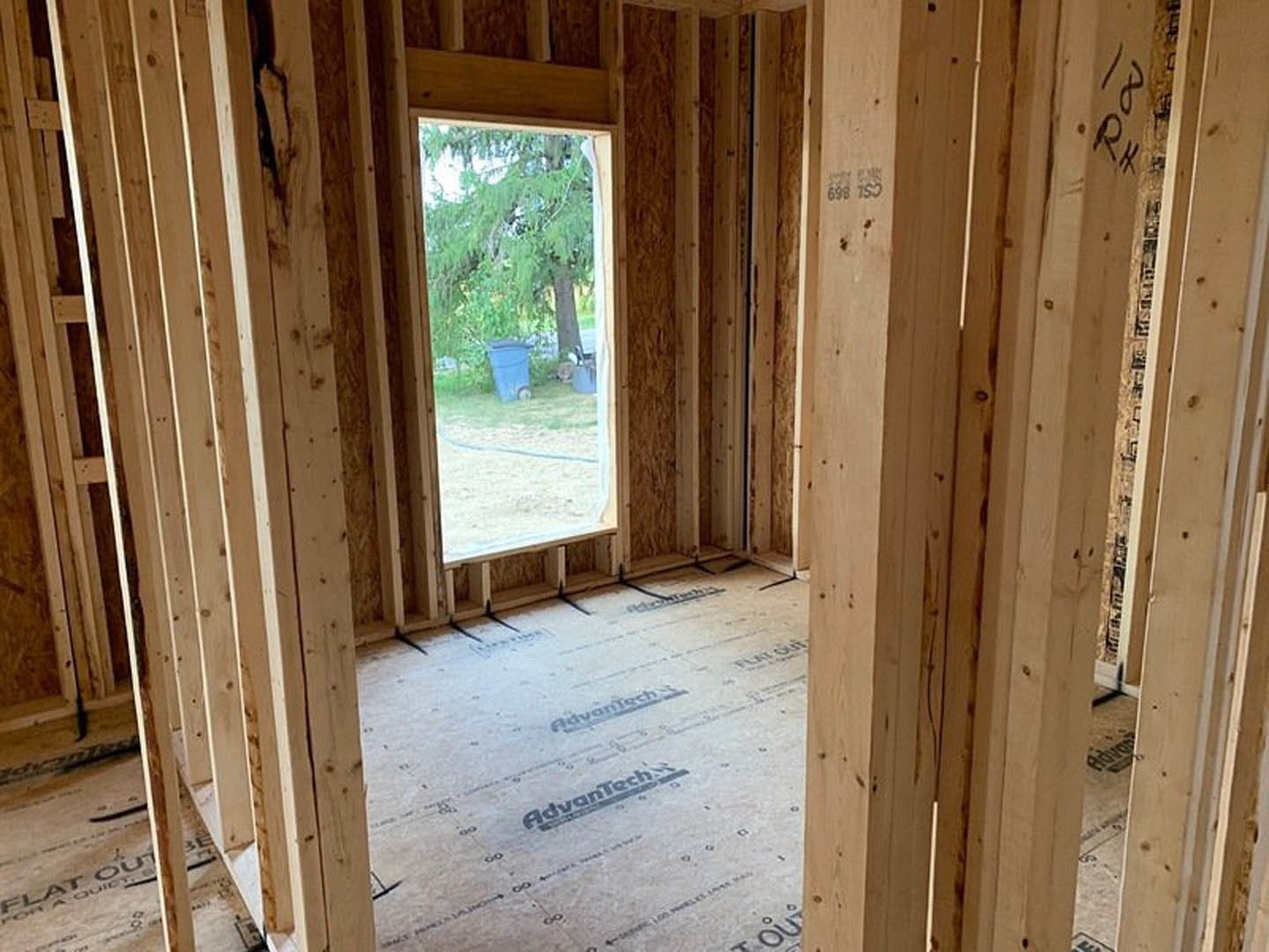 Room with light wood plank flooring, white walls, large window showing a tree and blue trash bin outside, simple white door on one side