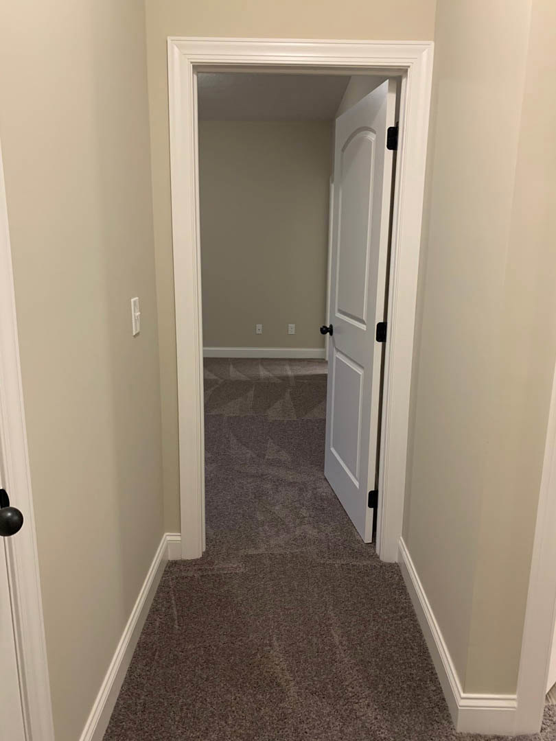 Hallway with beige carpet, white walls, open white door leading to adjacent room, chrome door handle, light switch visible on wall