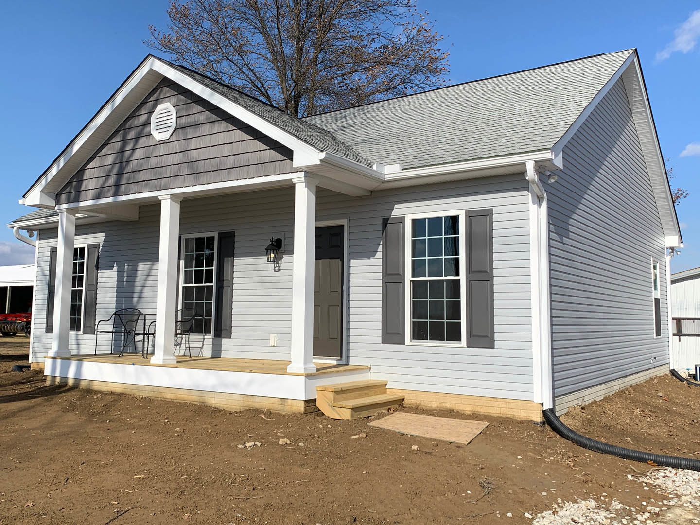 Gray siding house with white trim windows, covered porch featuring a chair, leafless tree in front yard, white vent on exterior wall, shingled roof under clear sky
