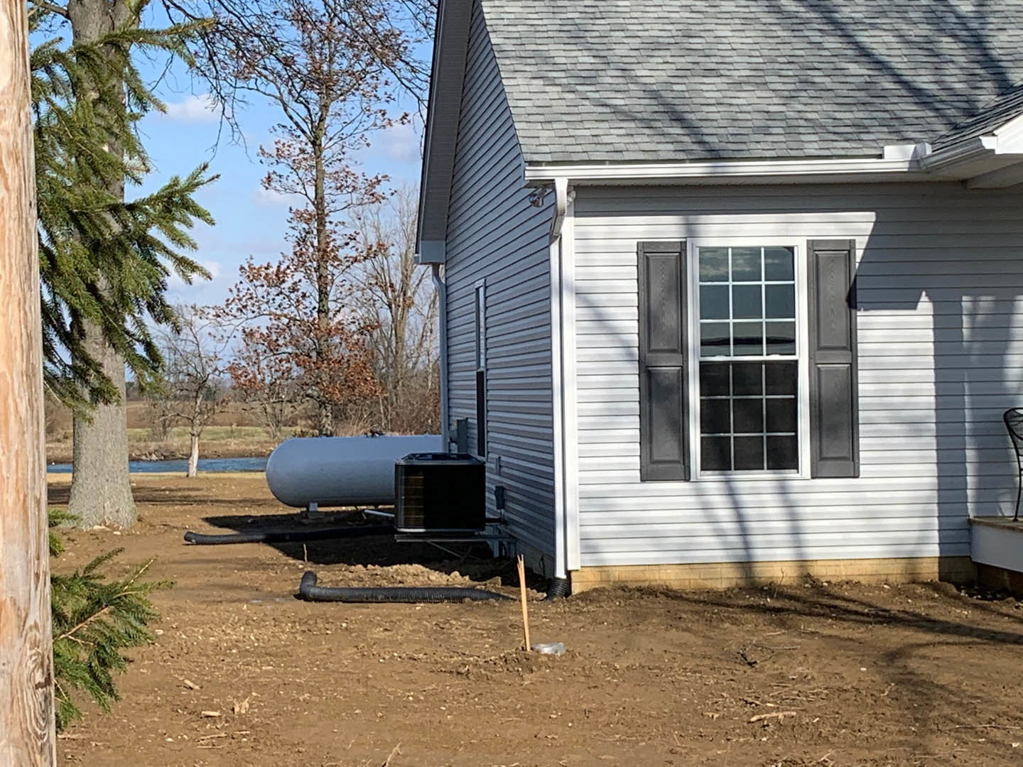 White-trimmed window with black shutter on light-colored siding, large cylindrical tank positioned beside the house, white pipe visible near foundation, surrounded by trees and