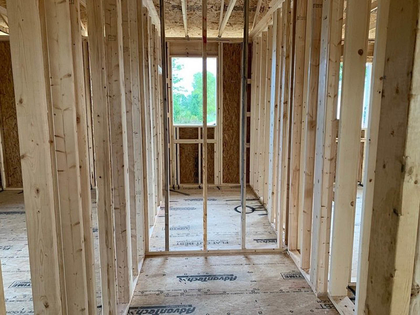 Wood-framed room under construction with exposed beams, unfinished floor, large window overlooking trees, and close-up of metal leg on floor