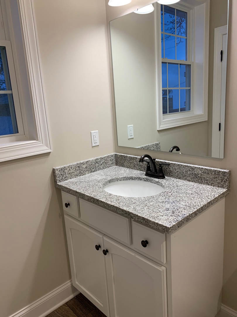 White ceramic vessel sink with matte black faucet on light stone countertop, rectangular mirror above, gray tile backsplash, window with blue sky, white cabinetry and drawers below