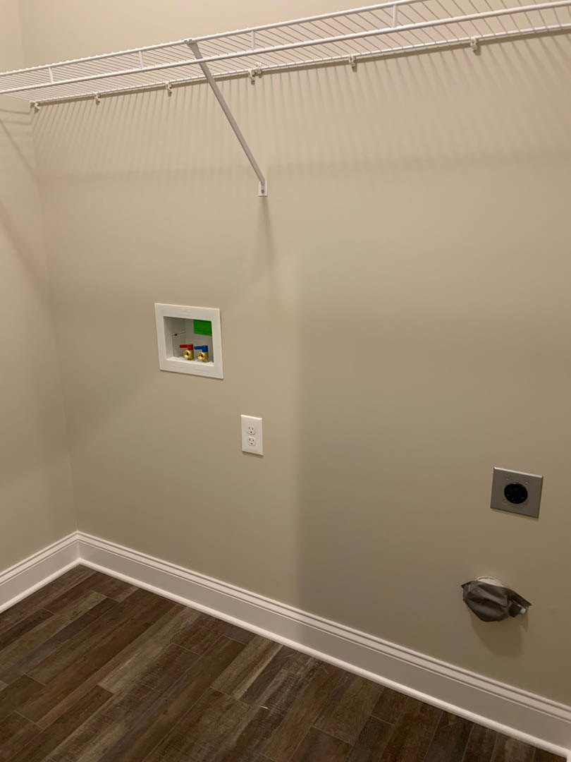 Open closet with wood flooring, clothes rack holding garments, white wall with black circular fixture, white shelf mounted above, and visible red and blue valve.