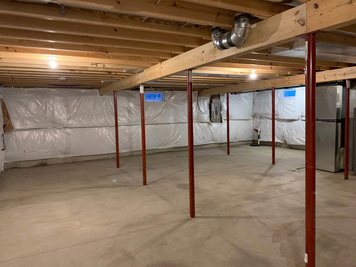 Basement with wooden ceiling beams, white painted wall, wooden floor, window, exposed concrete floor with red support poles, visible metal pipe secured with black rubber bands