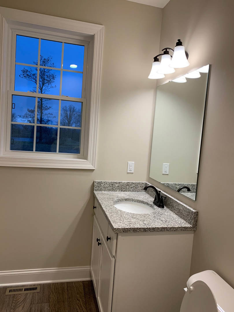 Modern bathroom featuring a stone countertop with a circular sink, chrome faucet, wall-mounted mirror, row of lights above, white toilet, and window overlooking a leafy tree.