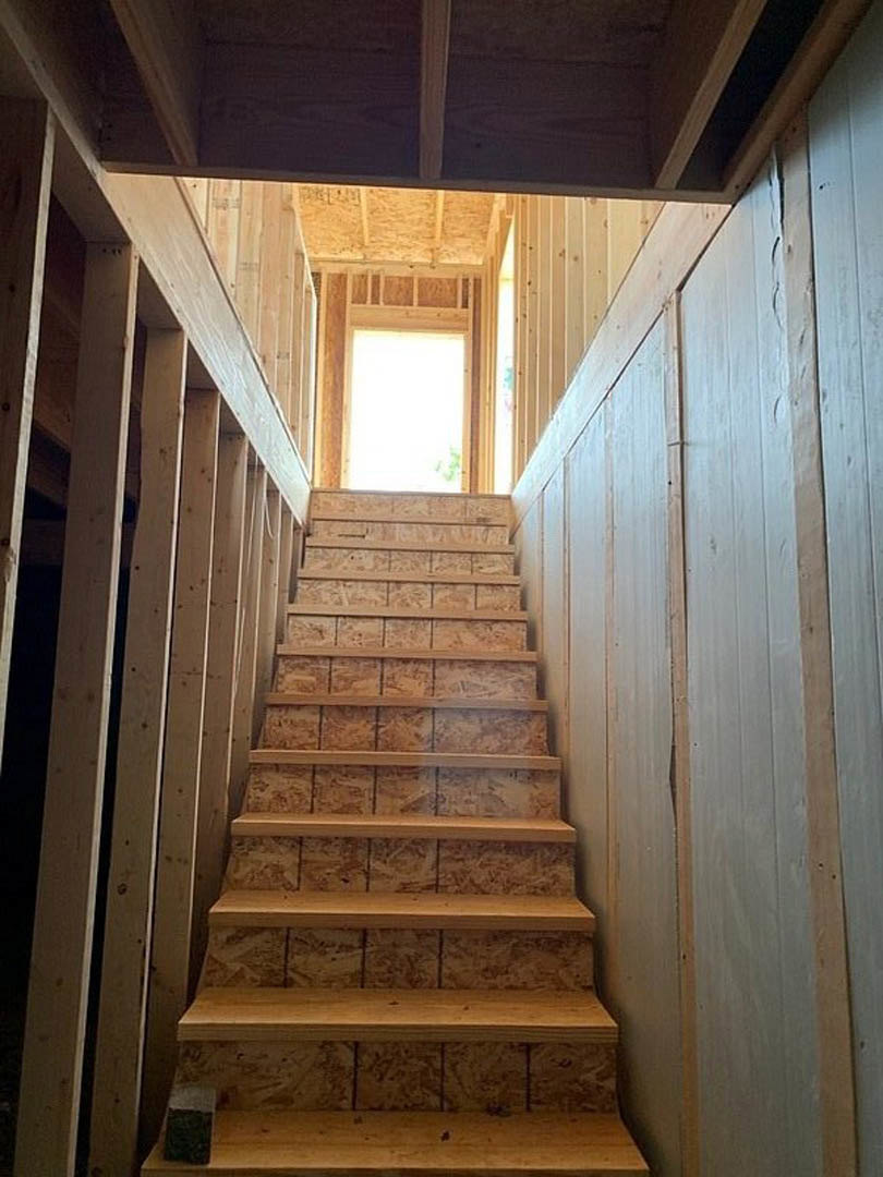 Wooden staircase with vertical plank wall, narrow handrail, and light wood flooring, natural light from nearby window, black square accent on white and brown surfaces