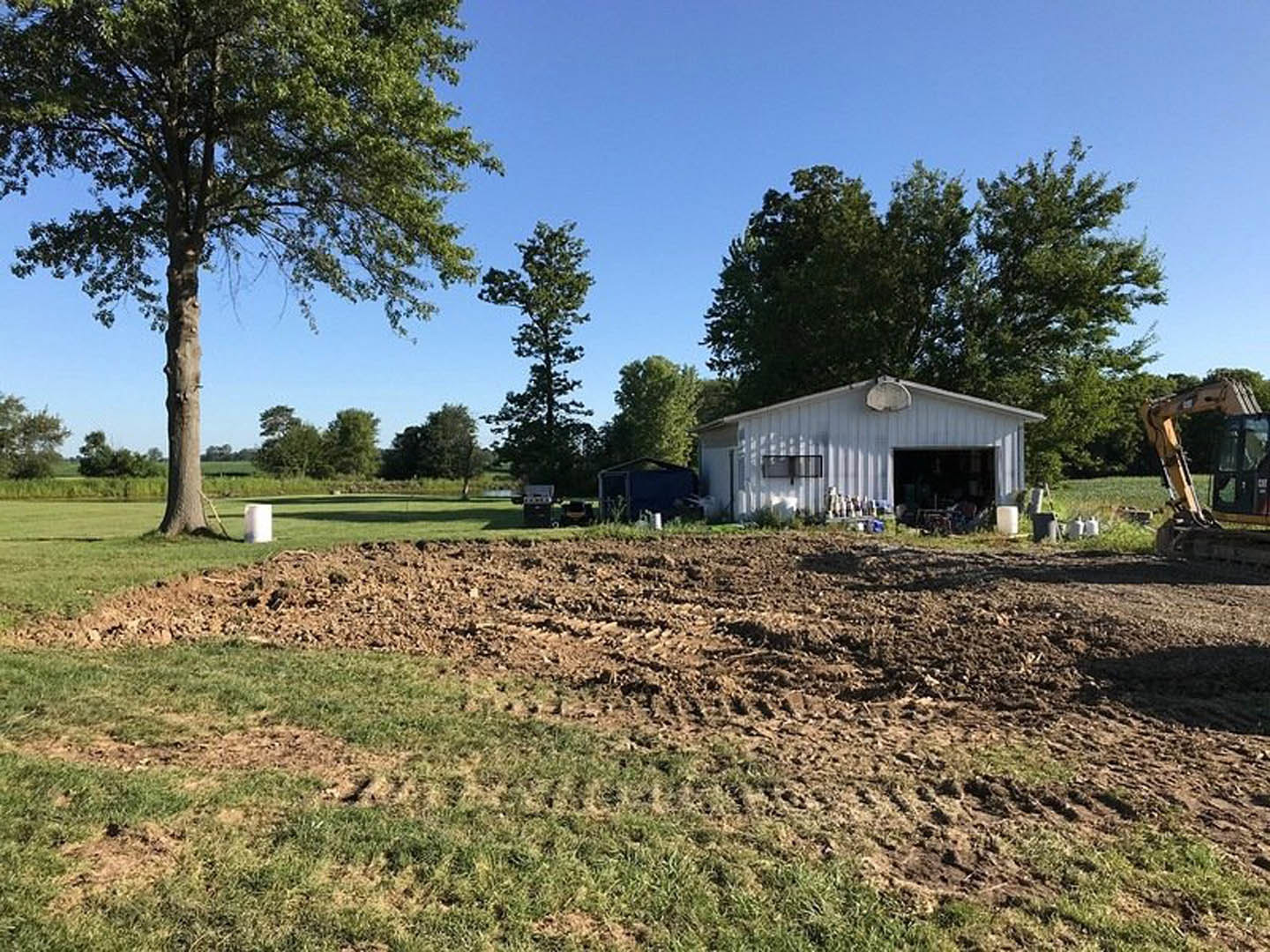White house with attached garage, basketball hoop in driveway, large dirt lot with tractor tracks, tree at edge of field, clear sky overhead.