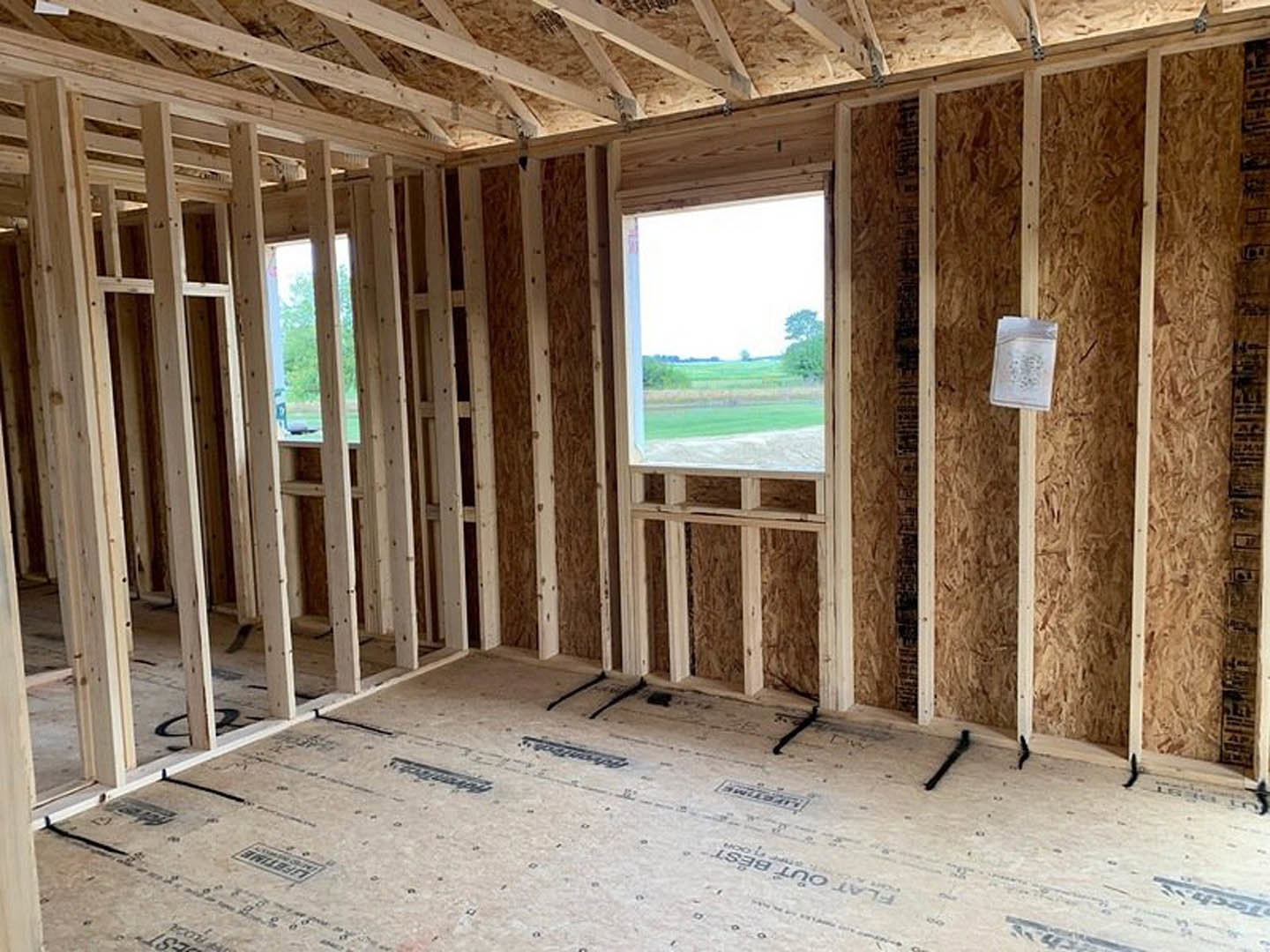 Unfinished room with exposed wood framing, visible wooden beams and planks, window overlooking grassy field, white insulation package with black text on floor