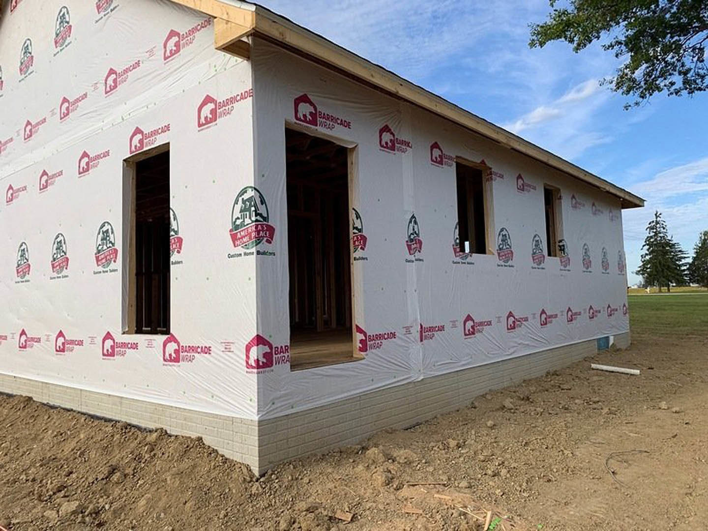 White exterior wall of a house under construction, partially covered with white plastic sheeting, tree branches with green leaves in foreground, unfinished window and door openings