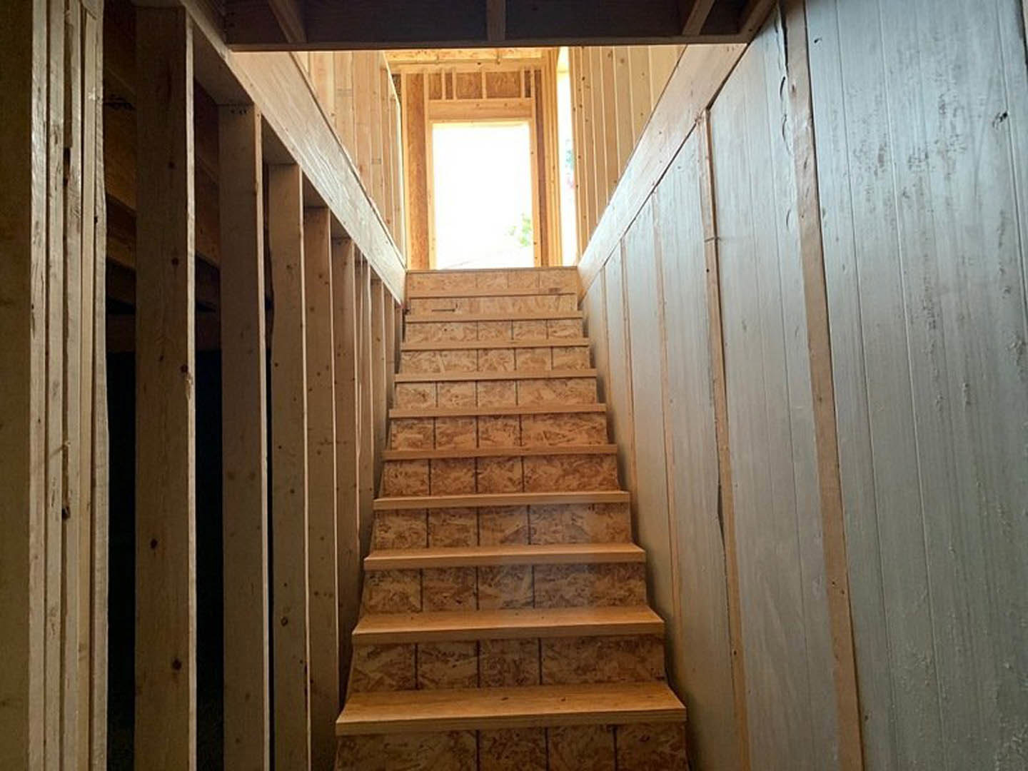 Wooden staircase with black handrail, adjacent to a white wall, sunlight streaming through a nearby window, and a wooden shelf mounted on the wall.