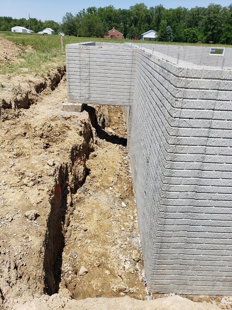 Concrete foundation wall with exposed soil and hole in the ground, surrounded by trees and outdoor landscape